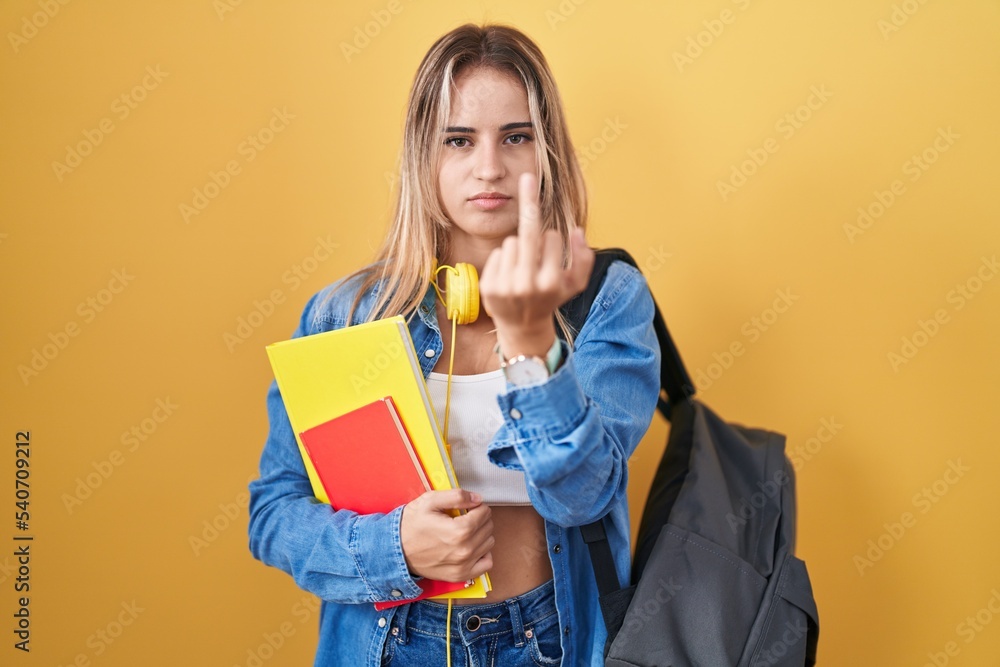 Young blonde woman wearing student backpack and holding books showing middle finger, impolite and rude fuck off expression Young blonde woman wearing student backpack and holding books showing middle finger, impolite and rude fuck off expression