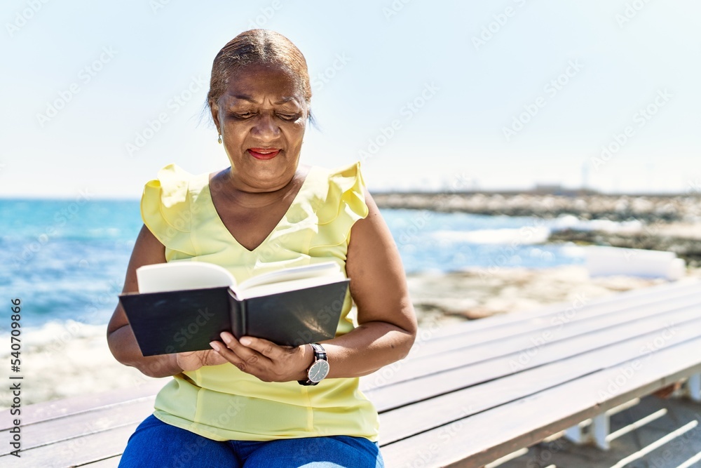 Senior african american woman reading book sitting on the bench at the beach.
