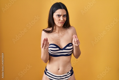 Young brunette woman wearing bikini over yellow background doing money gesture with hands, asking for salary payment, millionaire business