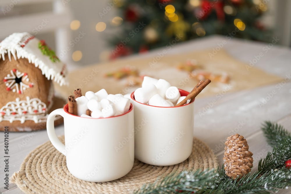 two white cup of cocoa with marshmallows and gingerbread house on table on background of Christmas tree two white cup of cocoa with marshmallows and gingerbread house on table on background of Christmas tree