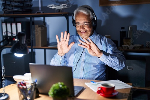 Hispanic senior man wearing call center agent headset at night showing and pointing up with fingers number nine while smiling confident and happy.