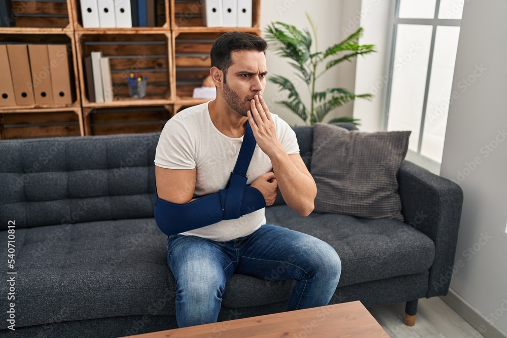 Young hispanic man with beard wearing arm on sling sitting at therapy consult covering mouth with hand, shocked and afraid for mistake. surprised expression Young hispanic man with beard wearing arm on sling sitting at therapy consult covering mouth with hand, shocked and afraid for mistake. surprised expression