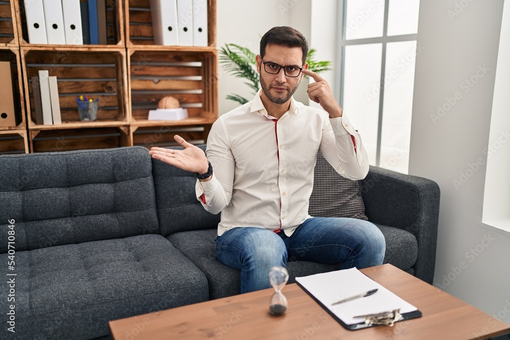 Young hispanic man with beard working at consultation office confused and annoyed with open palm showing copy space and pointing finger to forehead. think about it. Young hispanic man with beard working at consultation office confused and annoyed with open palm showing copy space and pointing finger to forehead. think about it.
