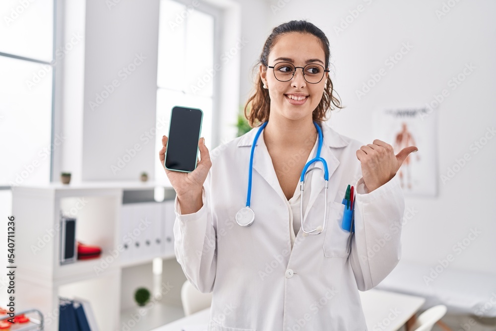 Young hispanic doctor woman holding smartphone showing screen pointing thumb up to the side smiling happy with open mouth Young hispanic doctor woman holding smartphone showing screen pointing thumb up to the side smiling happy with open mouth