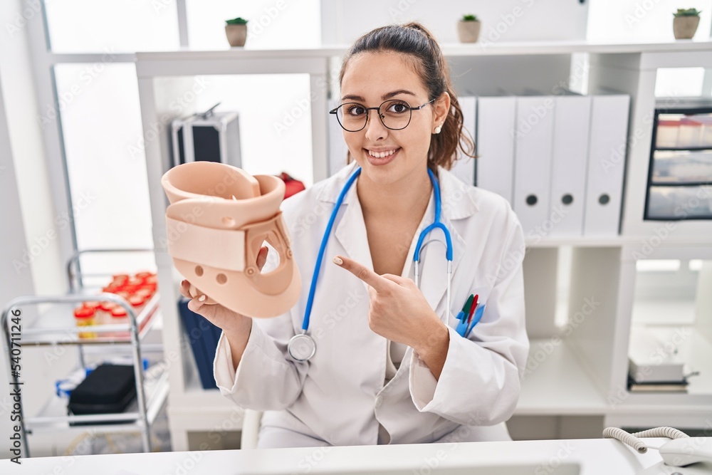 Young hispanic doctor woman holding cervical neck collar smiling happy pointing with hand and finger Young hispanic doctor woman holding cervical neck collar smiling happy pointing with hand and finger