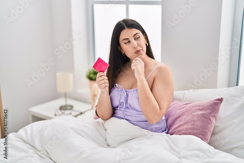 Young brunette woman wearing lingerie and holding condom on the bed serious face thinking about question with hand on chin, thoughtful about confusing idea