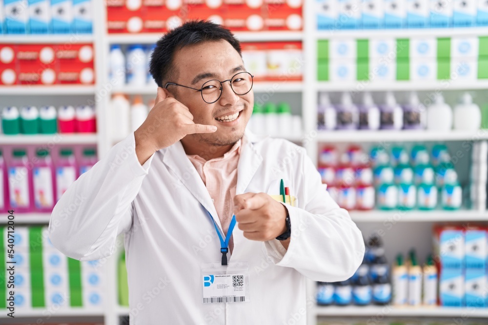 Chinese young man working at pharmacy drugstore smiling doing talking on the telephone gesture and pointing to you. call me.