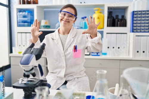 Hispanic girl with down syndrome working at scientist laboratory showing and pointing up with fingers number ten while smiling confident and happy.