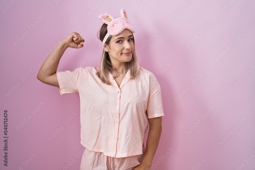 Blonde caucasian woman wearing sleep mask and pajama strong person showing arm muscle, confident and proud of power
