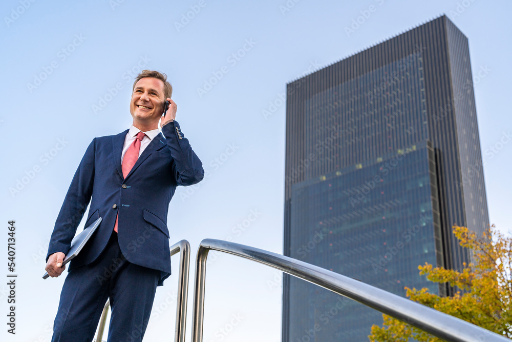 Happy businessman holding laptop talking on mobile phone by railing