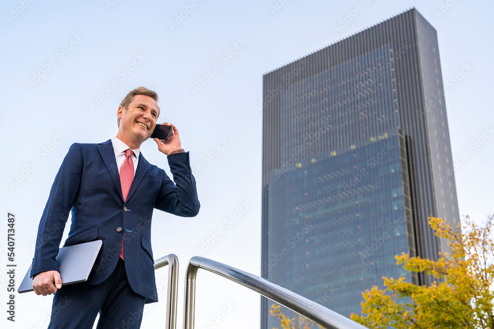 Happy mature businessman holding laptop talking on mobile phone by railing Happy mature businessman holding laptop talking on mobile phone by railing