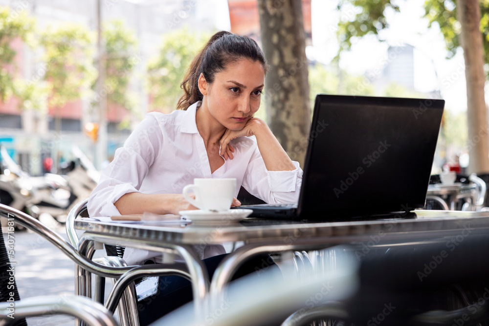 Businesswoman with hand on chin working on laptop at sidewalk cafe Businesswoman with hand on chin working on laptop at sidewalk cafe