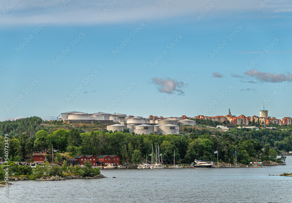 Sweden, Stockholm - July 16, 2022: Shoreline under Bergs oljehamn, oil port, in Nacka. Many oil tanks under blue morning sky. Historic red buildings and ships in small yacht port