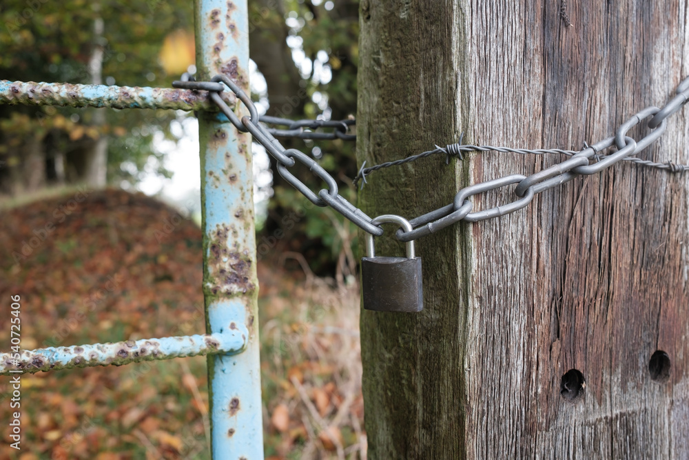 Padlock and chain linking an old metal gate and a wooden pole with barbed wire, blocking the passage to a private area. Padlock and chain linking an old metal gate and a wooden pole with barbed wire, blocking the passage to a private area.