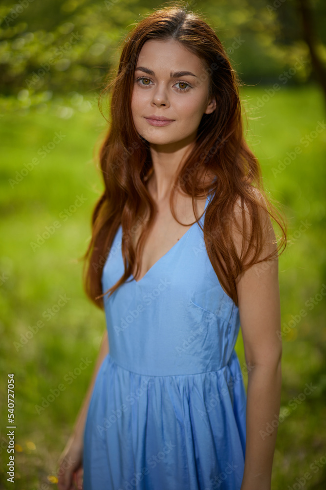 A beautiful woman in the springtime in nature stands by a tree in a blue dress, brooding sunset