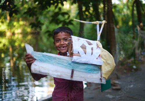 South asian little child standing with a handmade raft made with paper and sticks ,kid with toy in front of a pond