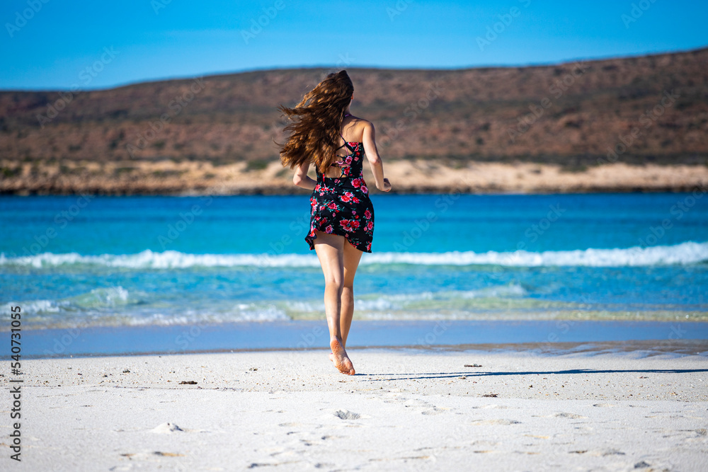 beautiful long-haired girl in a dress soaks her legs in turquoise water on a paradise beach at turquoise bay in cape range national park near exmouth, western australia; beach with red cliffs in the b