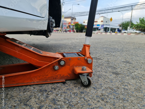 An orange jack is lifting the white car high to remove the wheels from the car. Repairing a car with damaged wheels such as a punctured car tire, a flat tire, or other accidents. 