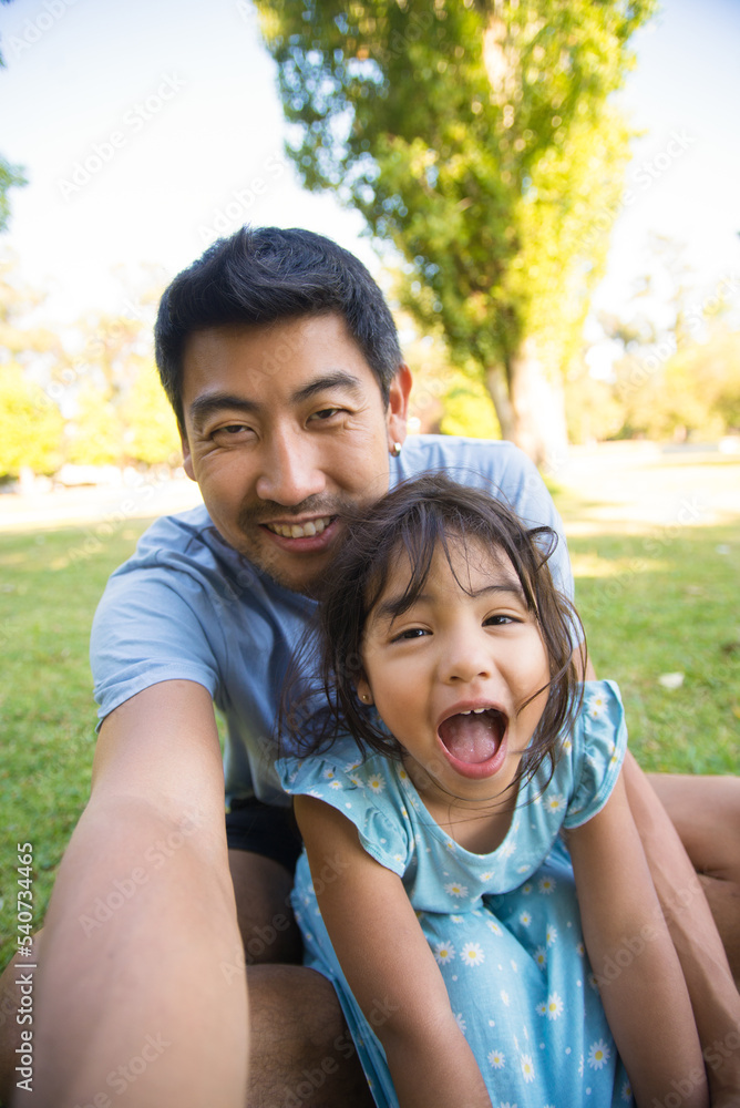 Asian dad taking selfie with his beautiful daughter. Handsome man spending vacation with his little girl in park both sitting on grass laughing looking at camera. Leisure, active rest outdoors concept Asian dad taking selfie with his beautiful daughter. Handsome man spending vacation with his little girl in park both sitting on grass laughing looking at camera. Leisure, active rest outdoors concept