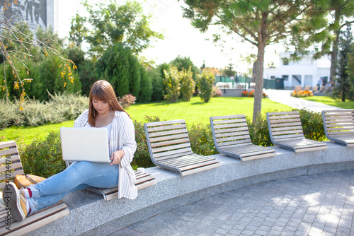 Young overwieght woman holding laptop and bagpack, dressed jeans and t-shirt siting in the syteet park  of the city photo