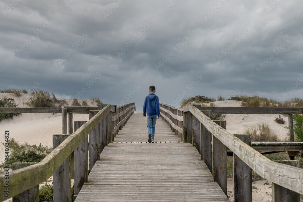 Boy walking on wooden deck to the sand on a day with very cloudy sky. High quality photo