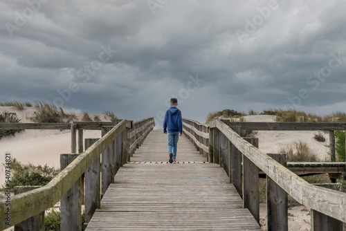 Boy walking on wooden deck to the sand on a day with very cloudy sky. High quality photo © amfer75