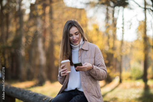 Young fashionable girl with smartphone and takeaway coffee in the park in autumn sits and smiles. Fashionable young woman in the autumn in the park writes sms or communicates online