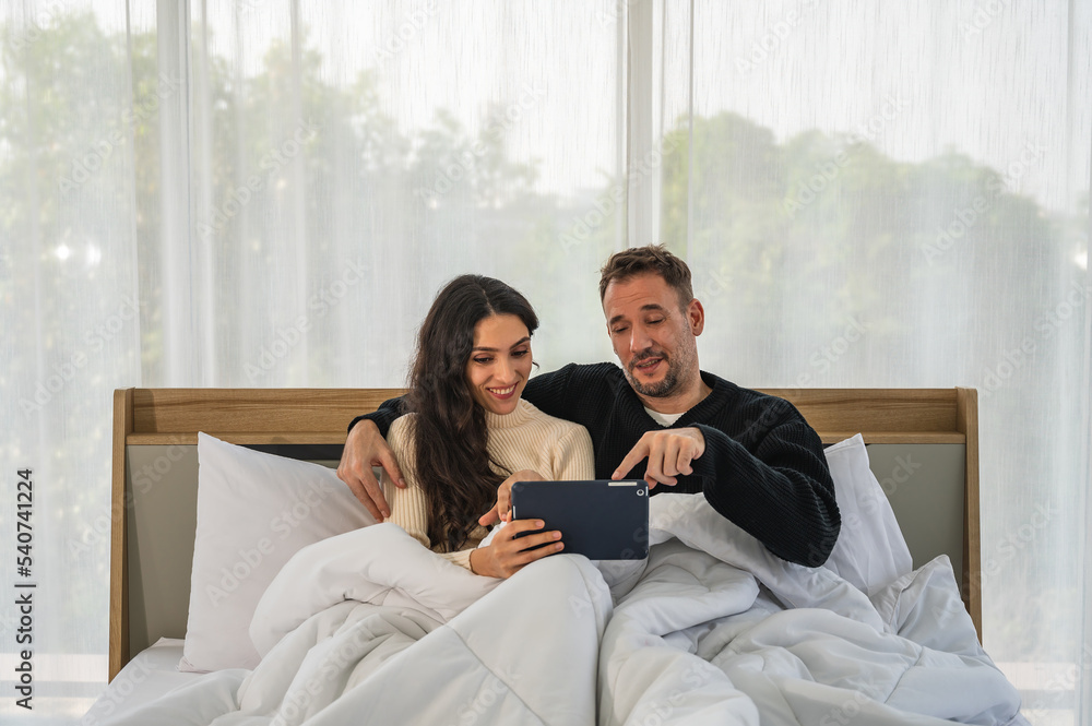 European couple man and woman smiling together looking at tablet mobile device sitting on bed under blanket. Male and female wearing warm winter clothing.