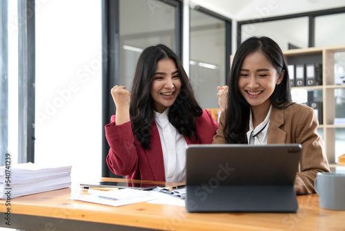 Two young Asian businesswomen show joyful expression of success at work smiling happily with a laptop computer in a modern office.