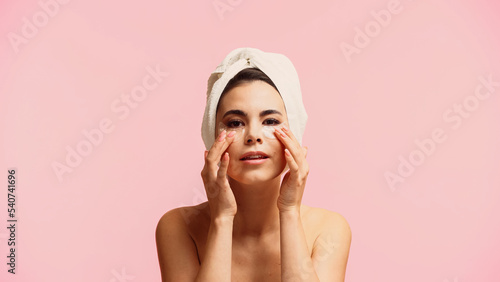 young woman with bare shoulders and towel on head applying moisturizing eye patches isolated on pink