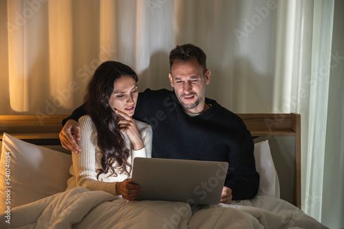 Caucasian couple sitting on their bed under blanket in dark room watching laptop screen looking serious.