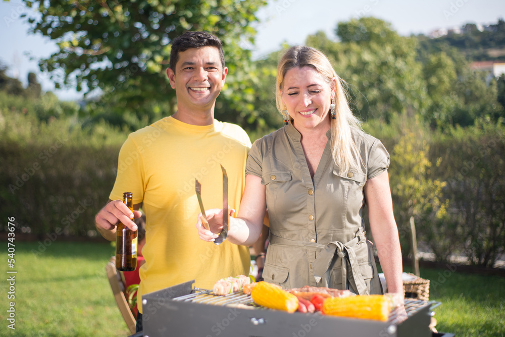 Happy husband and wife making BBQ in backyard. Dark-haired man in yellow T-shirt and woman in khaki dress standing near BBQ grid. Drinking beer. BBQ, cooking, food, family concept