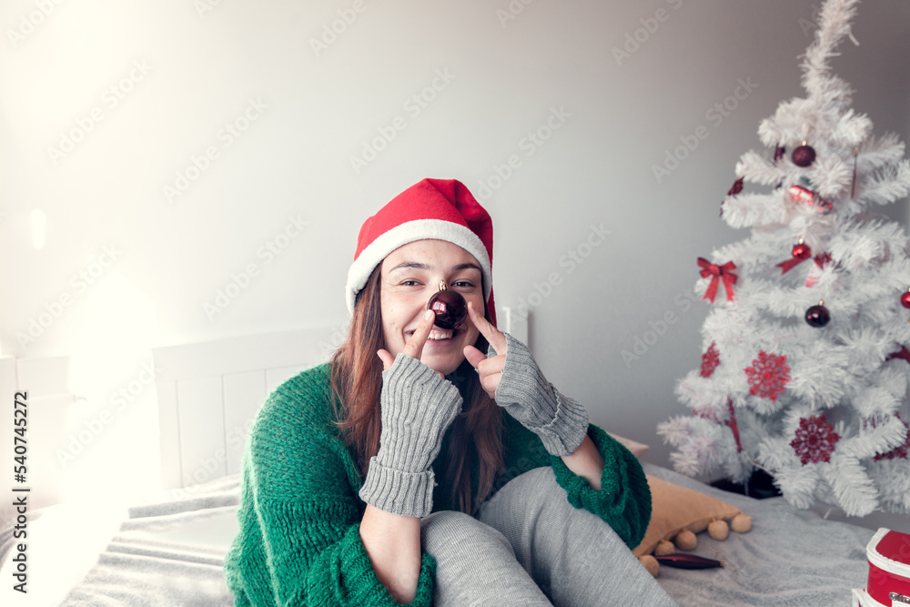 smiling girl sits on a bed against the background of a Christmas tree and holds a toy as a clown nose smiling girl sits on a bed against the background of a Christmas tree and holds a toy as a clown nose