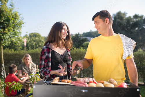 Caring father grilling meat and vegetables with daughter. Man in yellow T-shirt standing near BBQ grid looking at teenage girl. Wife and son in background. BBQ  cooking  food  family concept