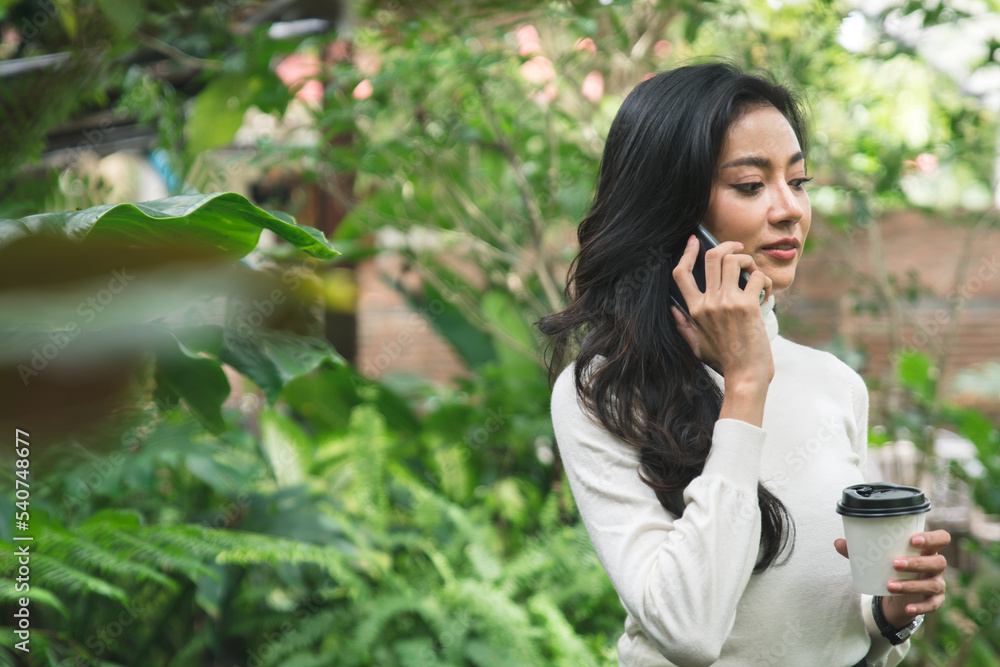 An Asian freelancer happily drinks coffee in the garden, she is using the phone to do business with customers. An Asian freelancer happily drinks coffee in the garden, she is using the phone to do business with customers.