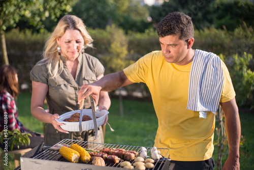 Happy husband and wife making BBQ in backyard on sunny day. Dark-haired man in yellow T-shirt and woman in khaki dress standing near BBQ grid. Putting food on plate. BBQ, cooking, food, family concept photo