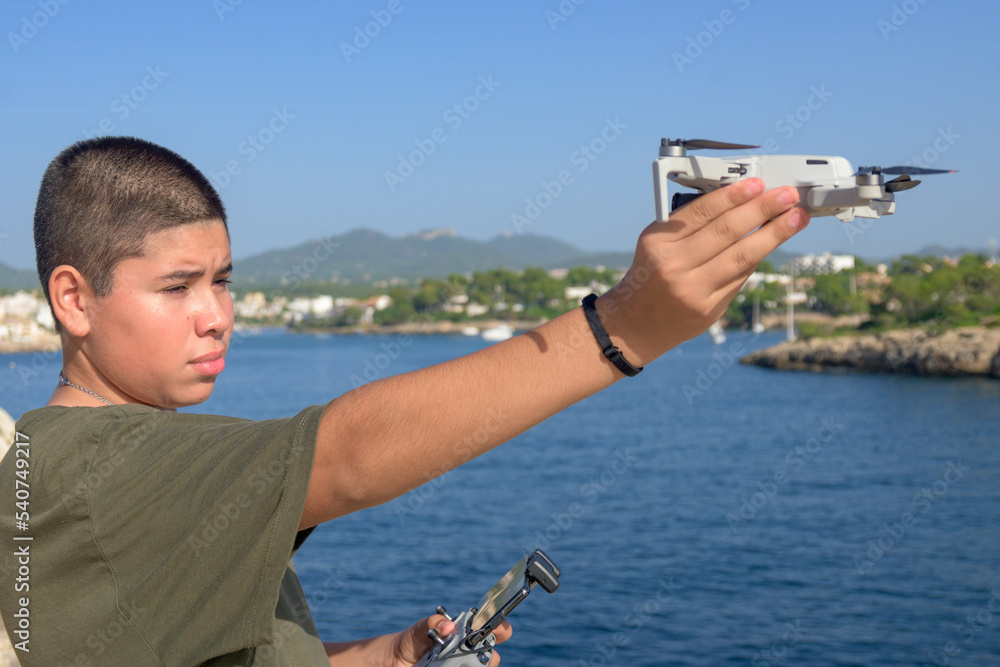 Naklejka premium portrait happy teenage boy, preparing and finalizing flight details for drone flight, Mediterranean sea background on a sunny day, technology concept Spain, Balearic Islands