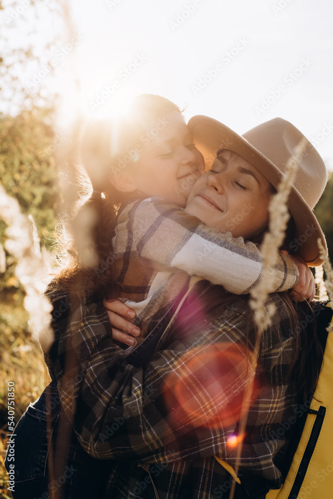 Obraz premium Vertical shot of loving pretty little daughter hugging with smiling young mother sit in grass on background beautiful autumn forest during epic sunset or sunrise. Concept family relations and travel