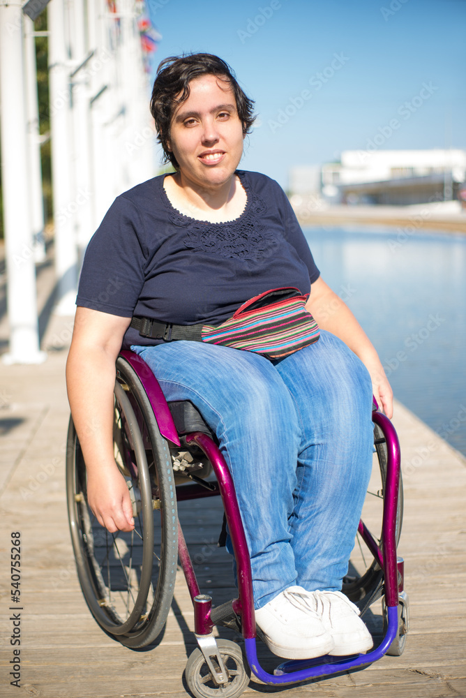 Portrait of relaxed woman in wheelchair. Caucasian woman in casual clothes on embankment, smiling at camera. Blue sky and flags in background. Portrait, beauty, happiness concept