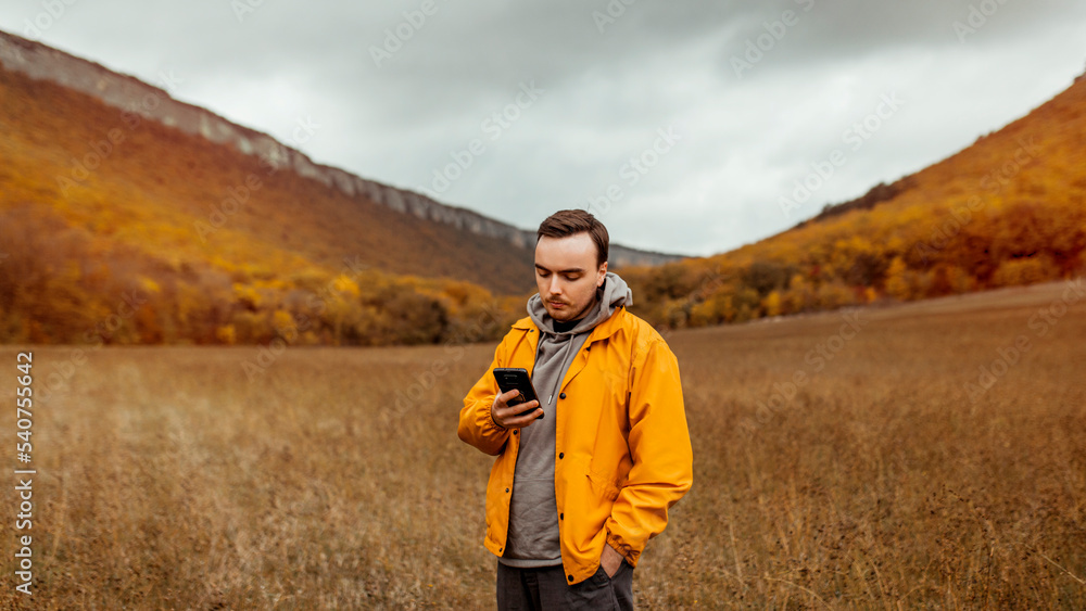 A young millennial male in a yellow jacket walks near the picturesque mountains and autumn forest in October and uses his smartphone A young millennial male in a yellow jacket walks near the picturesque mountains and autumn forest in October and uses his smartphone