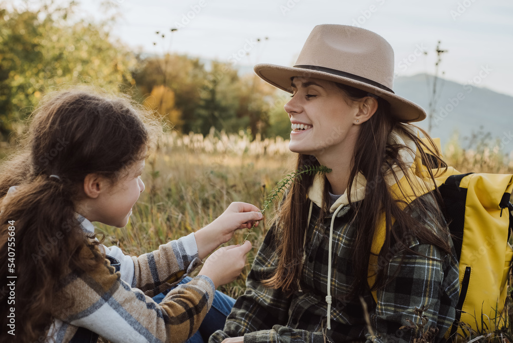 Cheerful young mother in hat and happy little daughter sitting in old grass enjoying weekend on background beautiful sunset or sunrise. Connection with nature and environmental protection concept