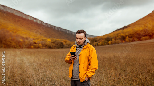 A young millennial male in a yellow jacket walks near the picturesque mountains and autumn forest in October and uses his smartphone