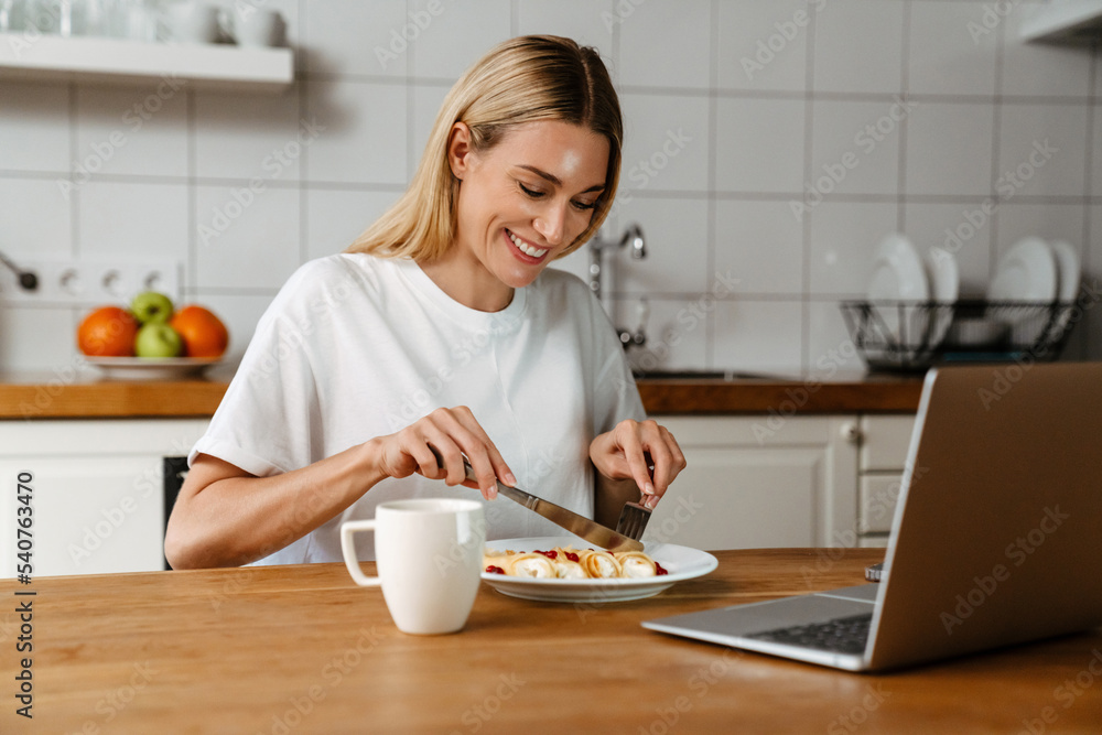Happy woman smiling and using laptop while having breakfast at home Happy woman smiling and using laptop while having breakfast at home