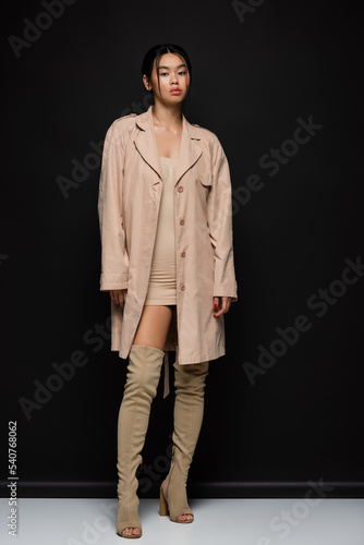 Stylish asian woman in trench coat and boots standing on black background