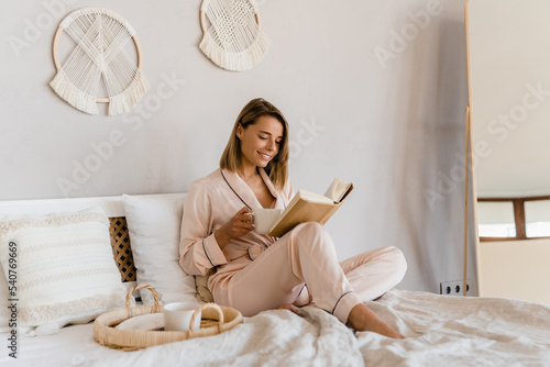 pretty smiling woman relaxing at home on bed in morning in pajamas reading book