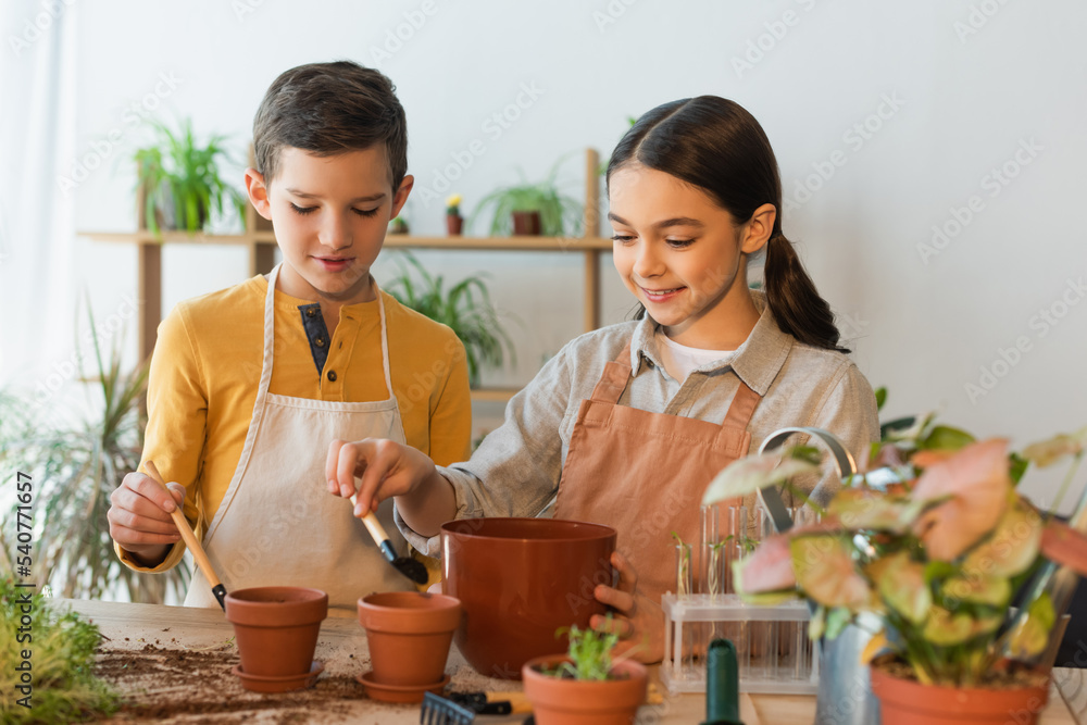 Smiling girl pouring ground in flowerpot near friend and plants at home. Smiling girl pouring ground in flowerpot near friend and plants at home.