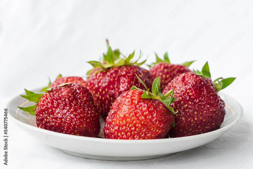 Red ripe strawberry in the white plate, light background. Plate with strawberries on white fabric. Summer vitamin berries juicy strawberry with strawberry leaf. Healthy eating and food concept Red ripe strawberry in the white plate, light background. Plate with strawberries on white fabric. Summer vitamin berries juicy strawberry with strawberry leaf. Healthy eating and food concept
