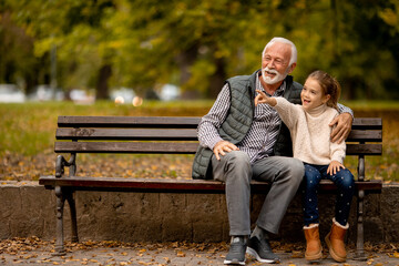 Grandfather spending time with his granddaughter on bench in park on autumn day