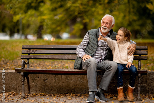 Fototapeta Grandfather spending time with his granddaughter on bench in park on autumn day