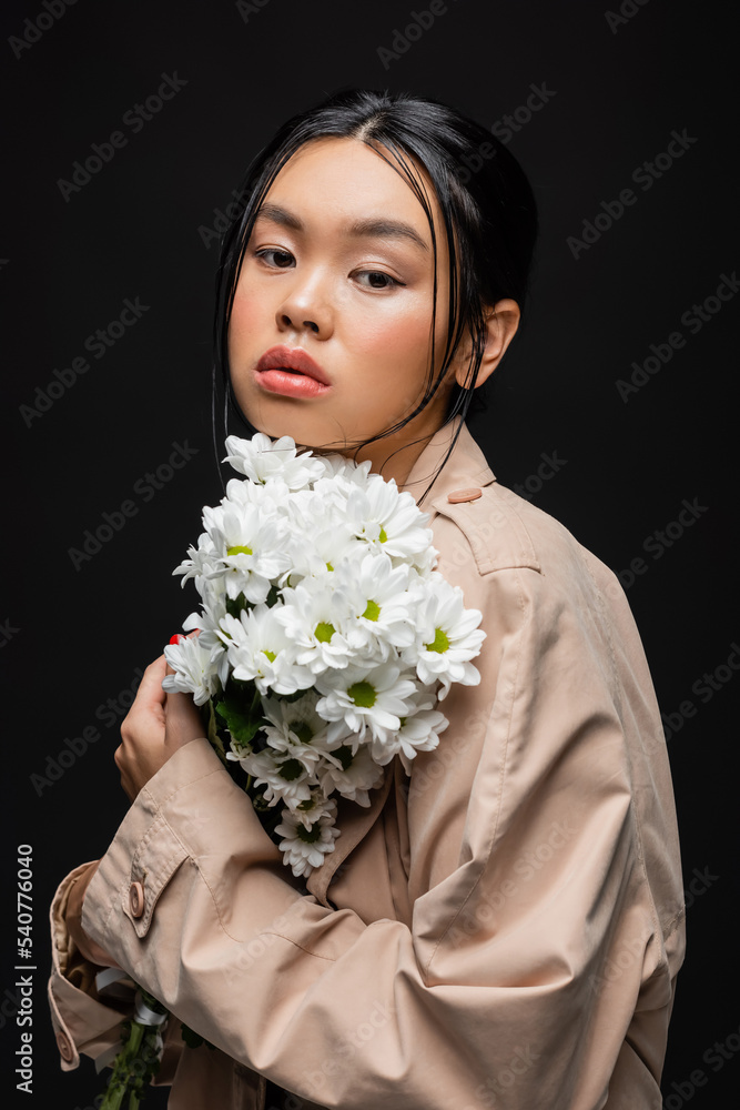 Stylish asian woman in trench coat holding white chrysanthemums isolated on black Stylish asian woman in trench coat holding white chrysanthemums isolated on black